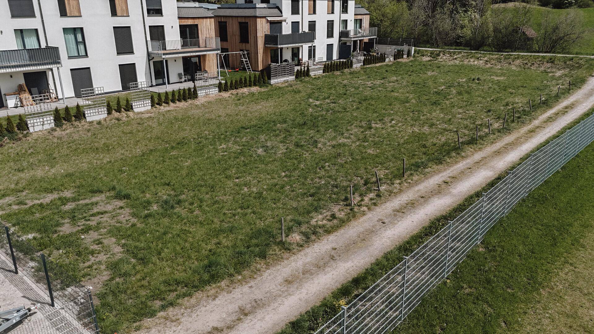 Weitläufiges grünes Grundstück mit modernen Gebäuden auf der linken Seite und einem Feldweg mit Zaun rechts.