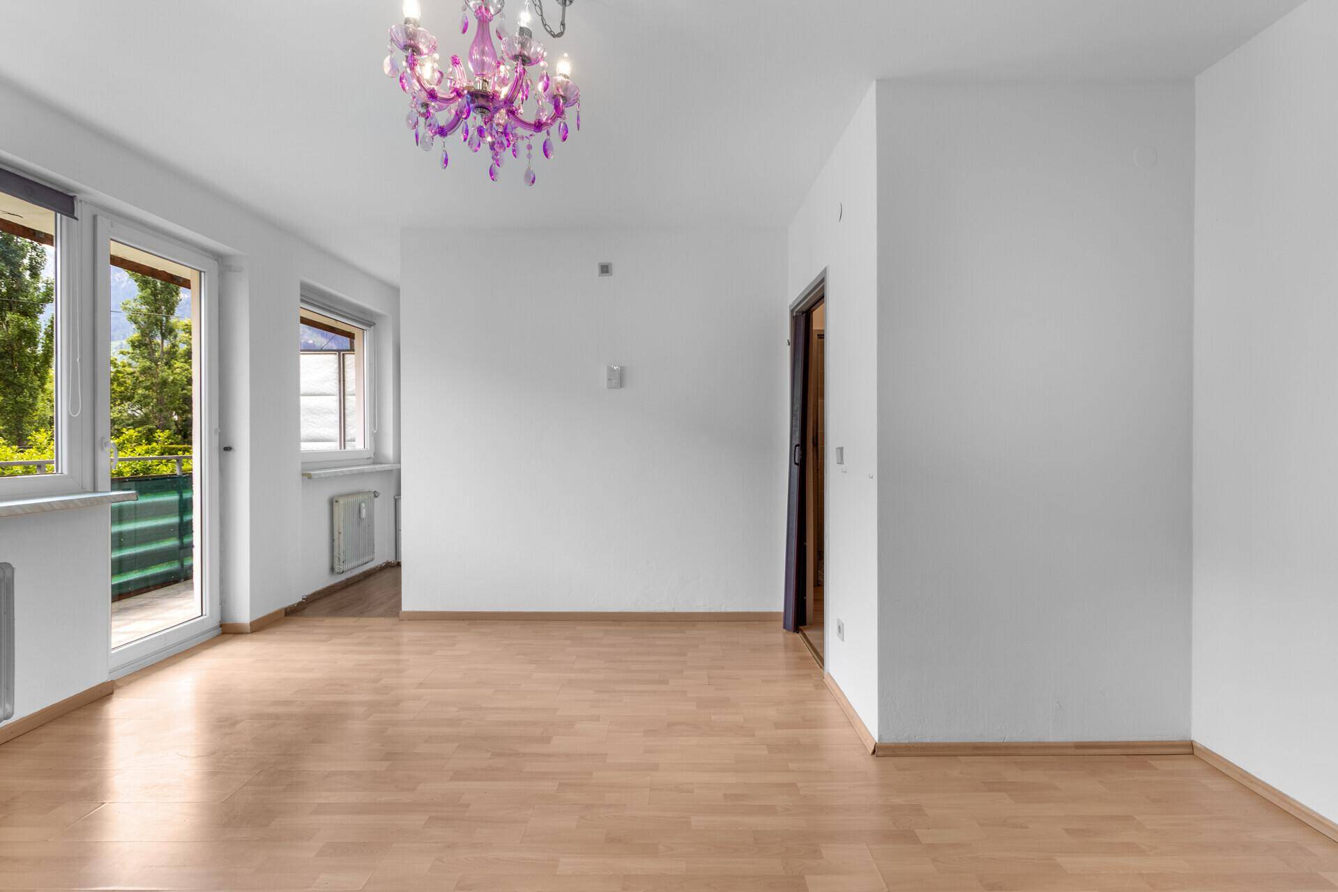 Geräumiges Zimmer mit hellem Holzboden, Zugang zum Balkon und moderner Deckenleuchte.