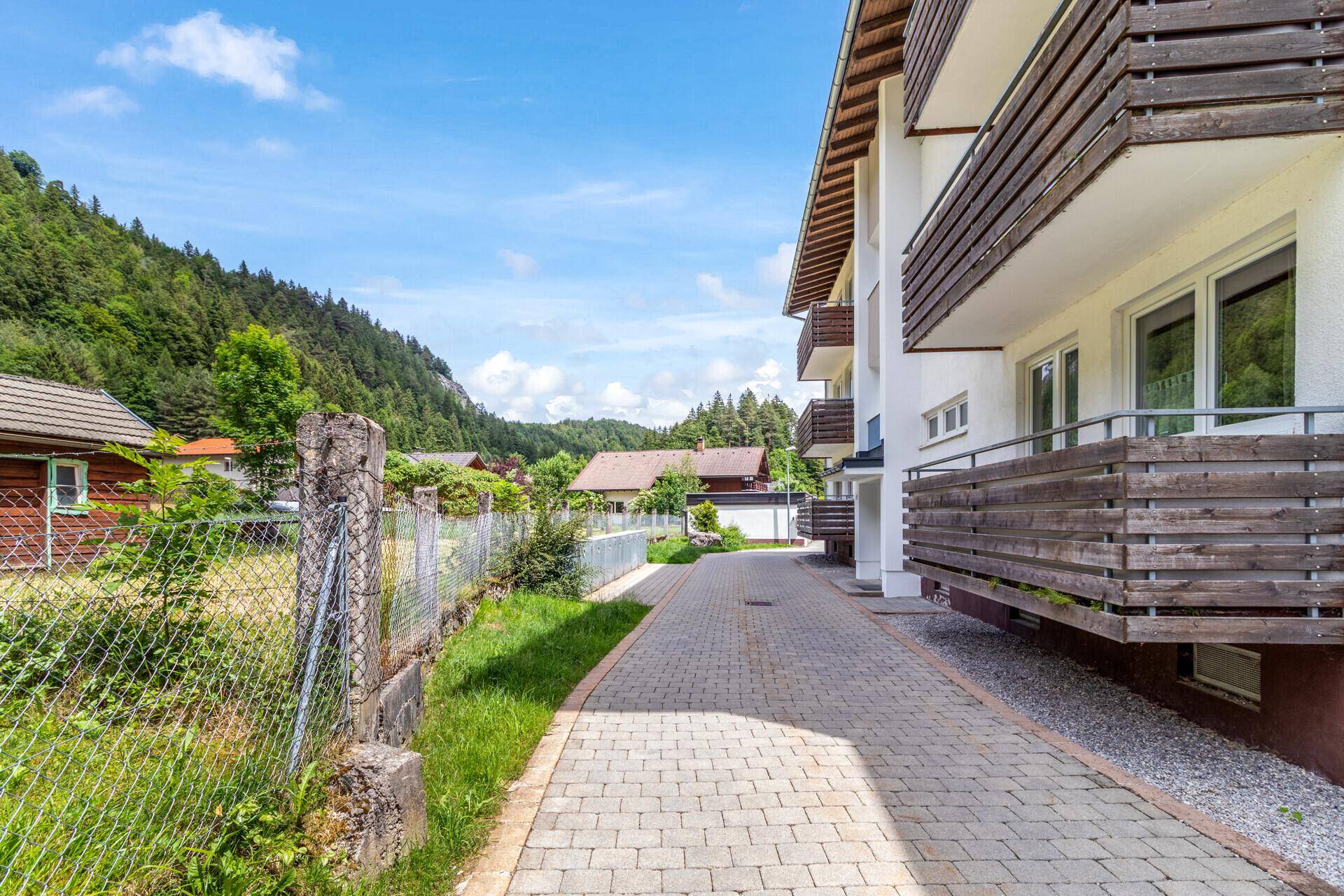 Außenansicht des Gebäudes mit gepflasterter Zufahrt und Blick auf die bewaldeten Berge.