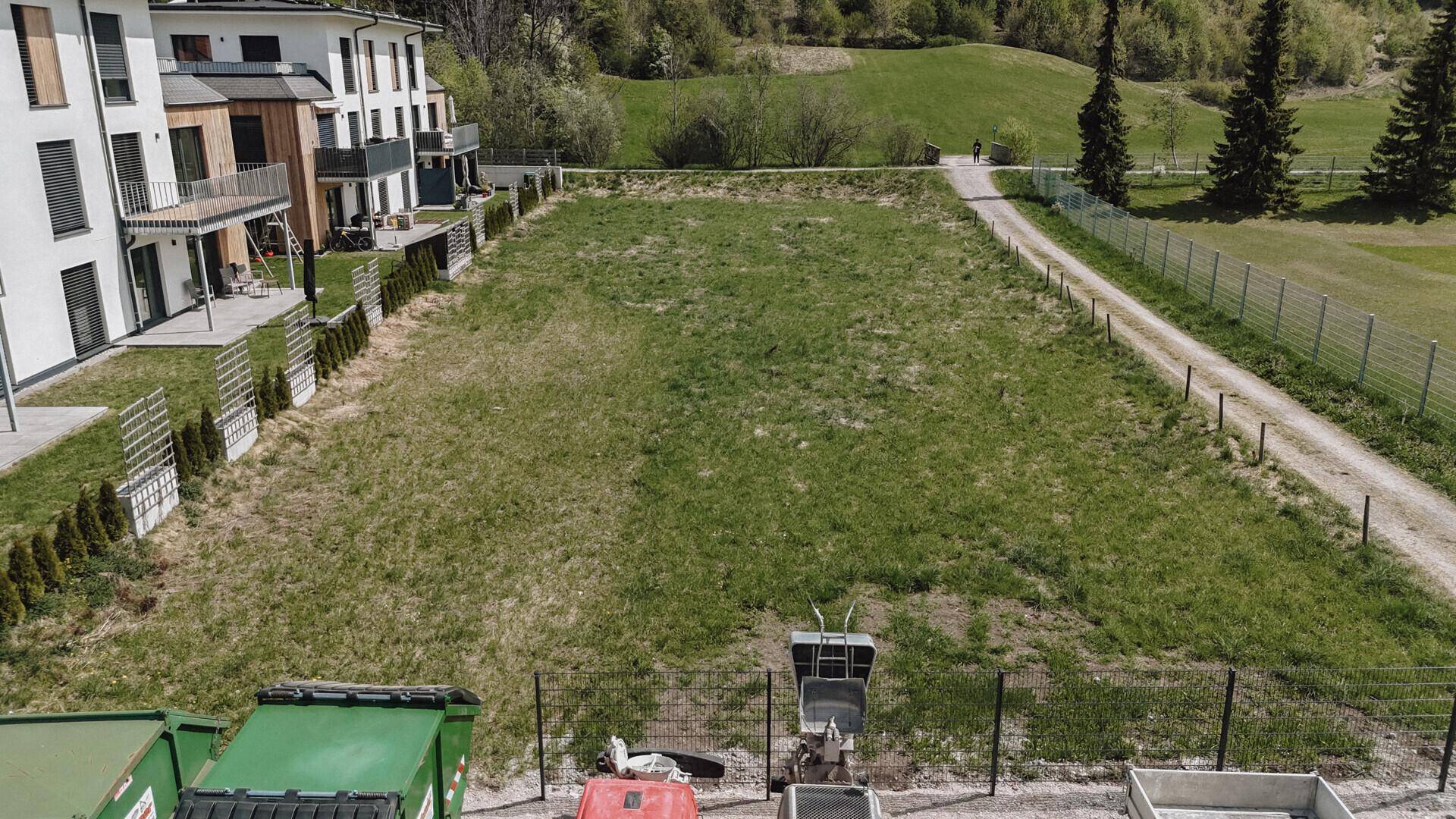 Großes, grünes Grundstück mit modernen Gebäuden links und einem Feldweg mit Zaun rechts, im Vordergrund Baugeräte.