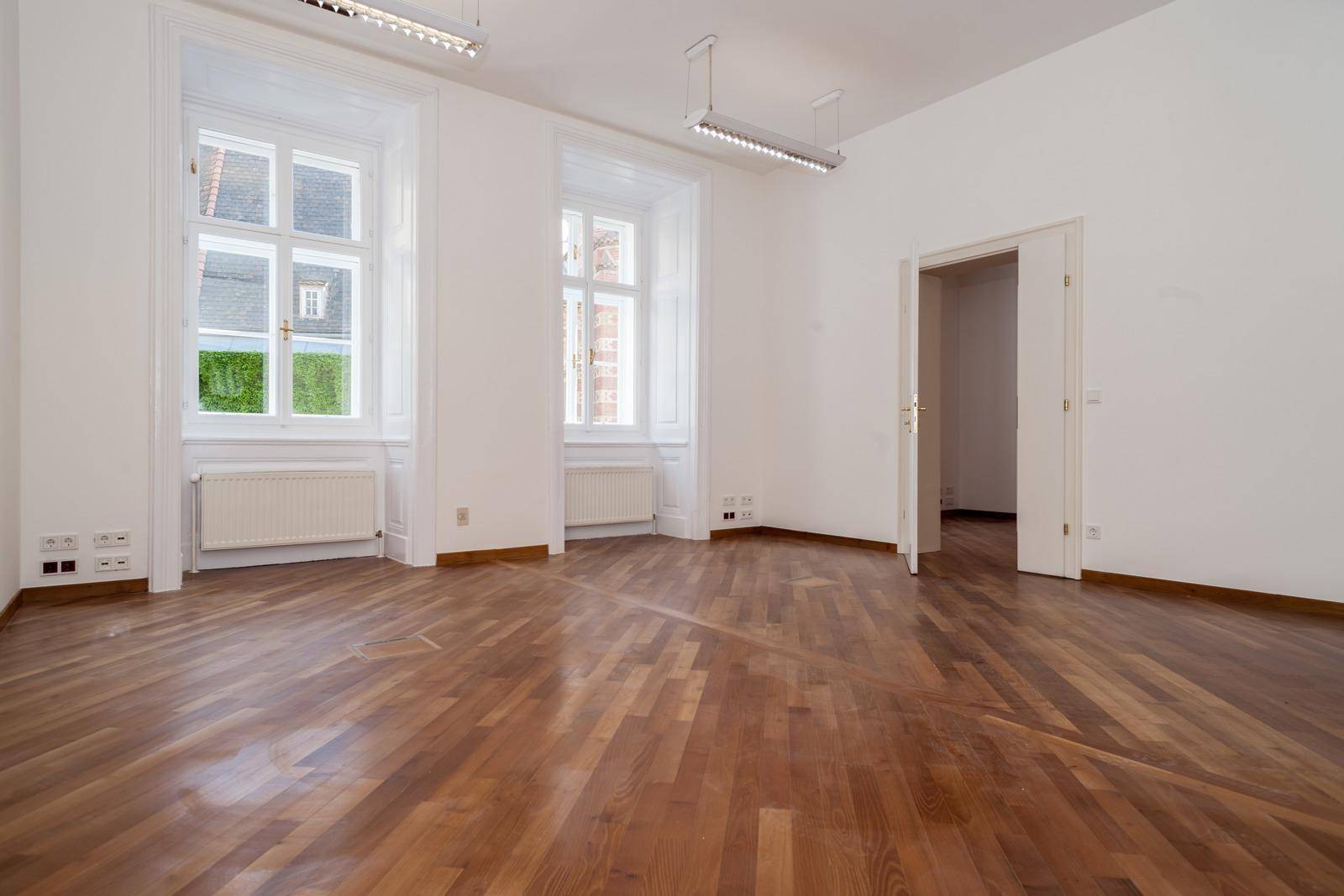 Heller Büroraum mit Fischgrätparkett und großen Fenstern, die viel Tageslicht hereinlassen.