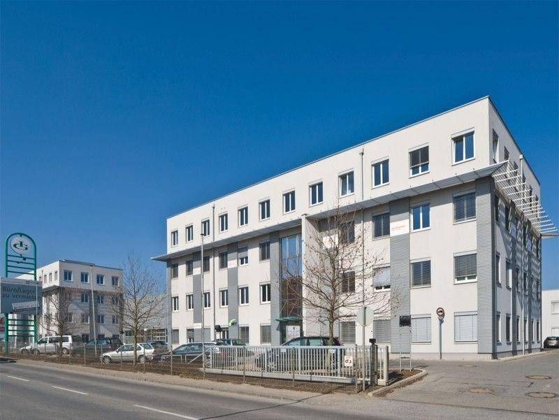 Modernes Bürogebäude mit mehreren Etagen und großen Fenstern, umgeben von Parkplätzen.
