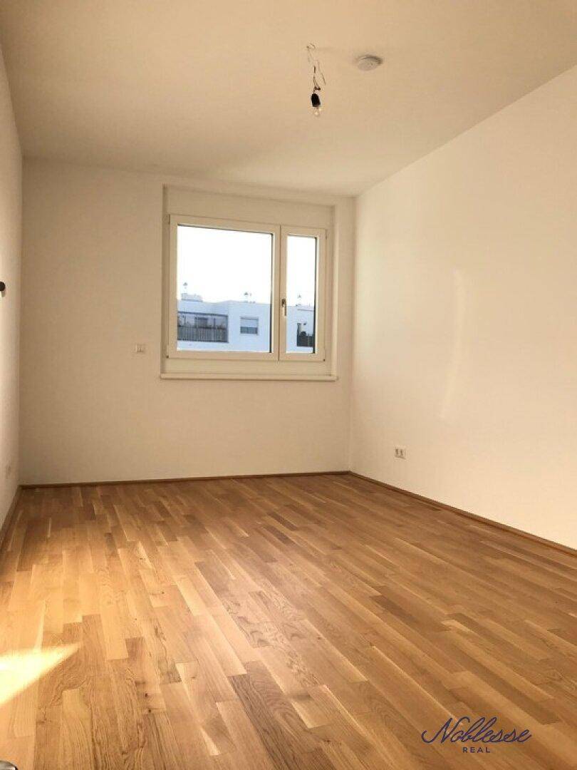 Leeres Schlafzimmer mit Holzboden und einem großen Fenster, das viel Licht hereinlässt.