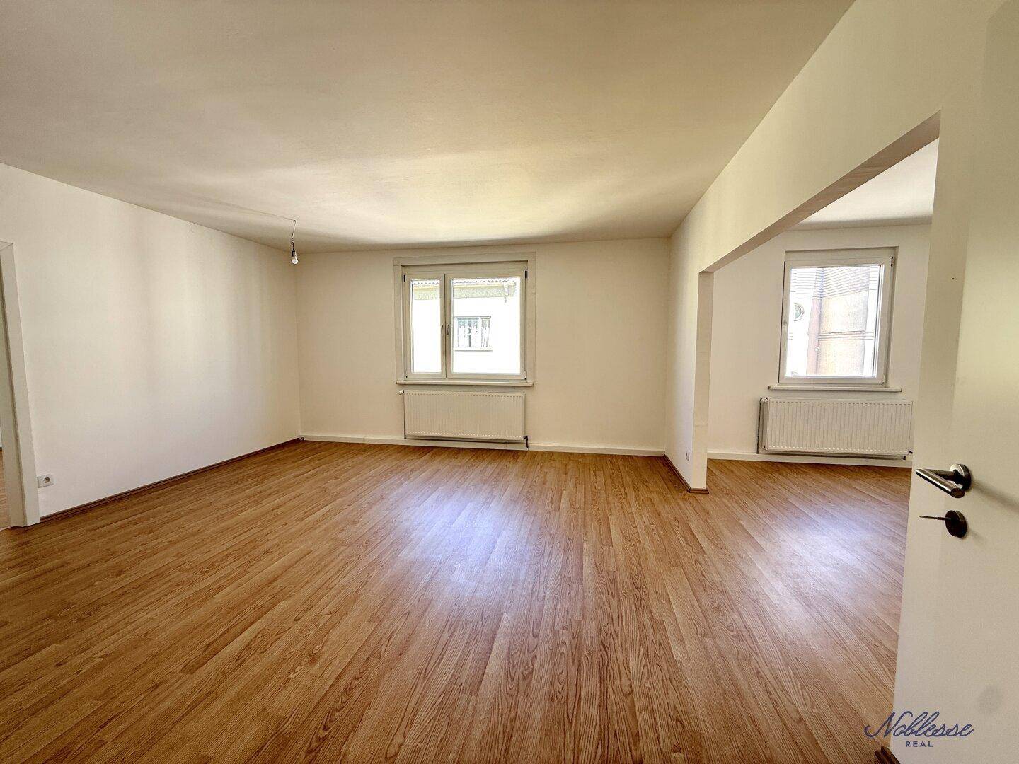 Heller Wohnbereich mit Laminatboden und zwei Fenstern, bietet viel Potenzial für eine gemütliche Einrichtung.