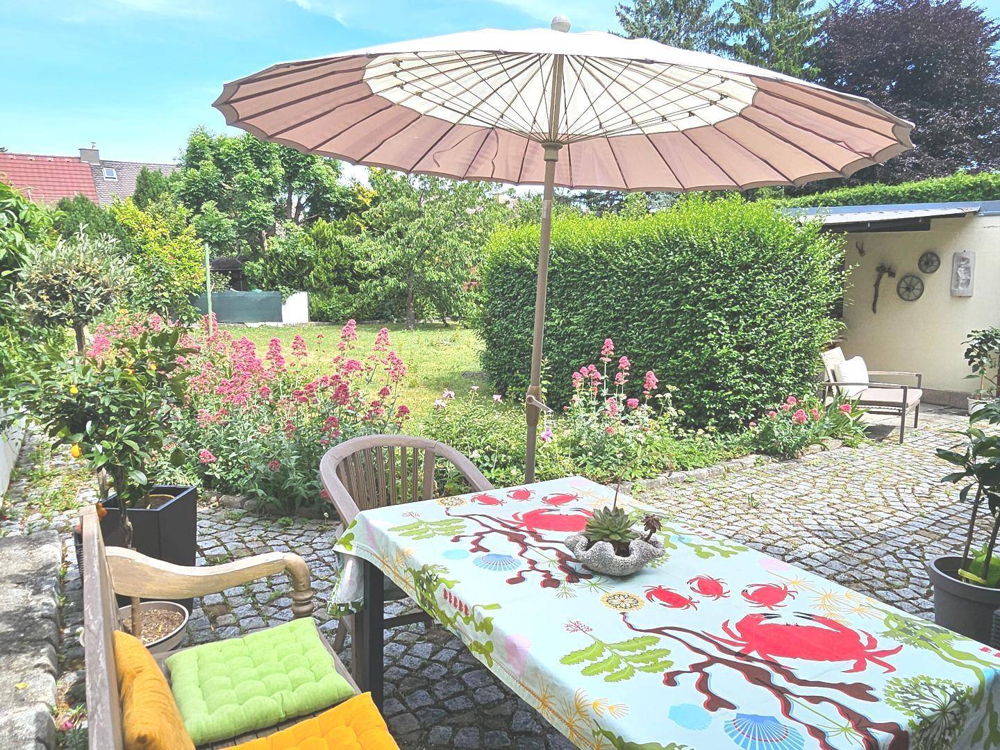 Sonniger Gartenbereich mit Sitzgelegenheiten, Sonnenschirm und blühenden Pflanzen.