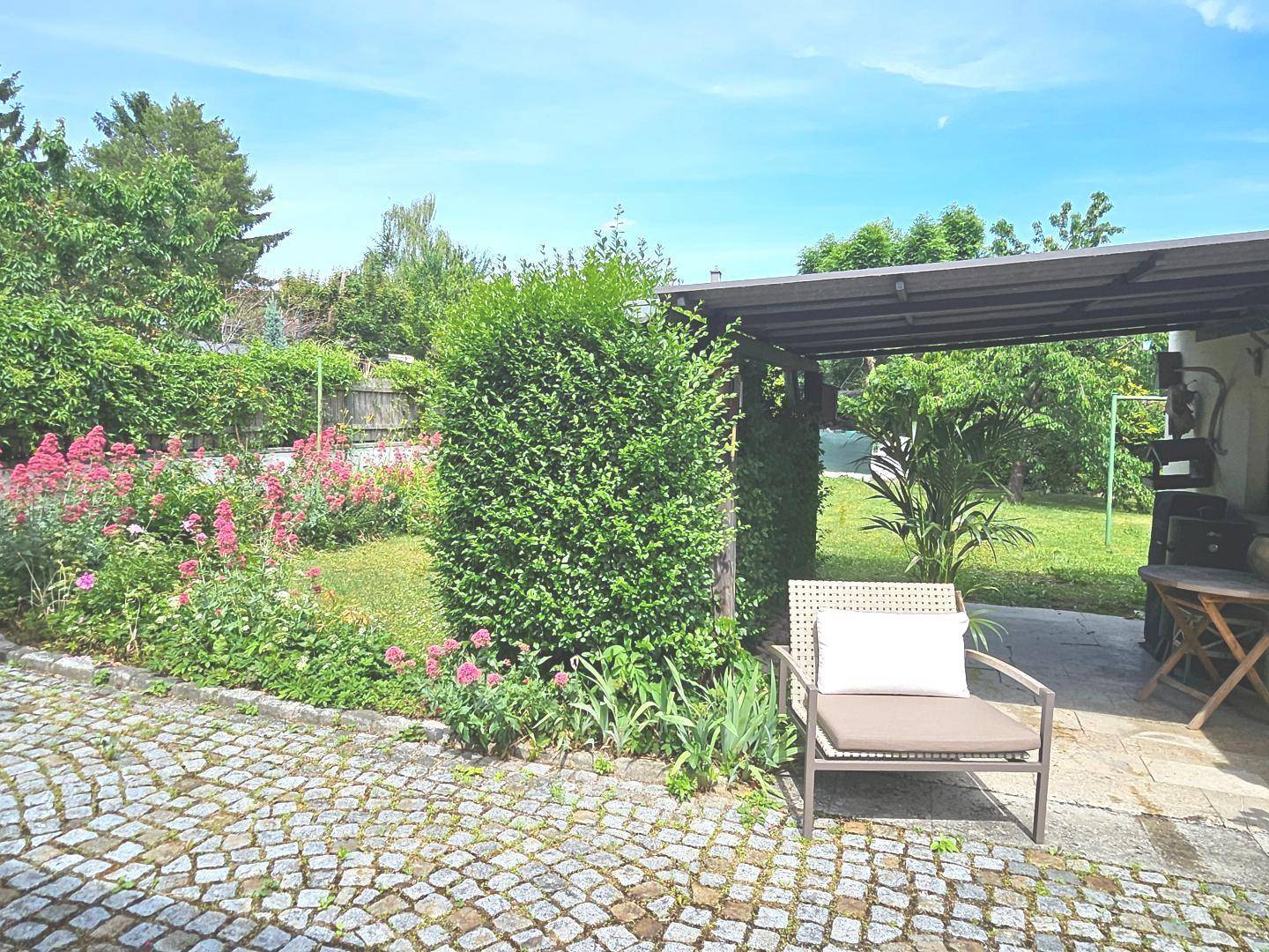Gepflasterter Gartenbereich mit Sitzgelegenheit, umgeben von Grünflächen und Blumen.