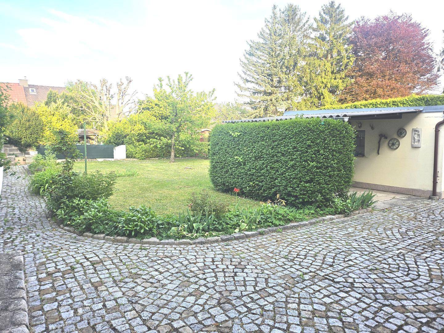 Weitläufiger Garten mit Rasenfläche, Hecken und Bäumen, umgeben von einer gepflasterten Auffahrt.