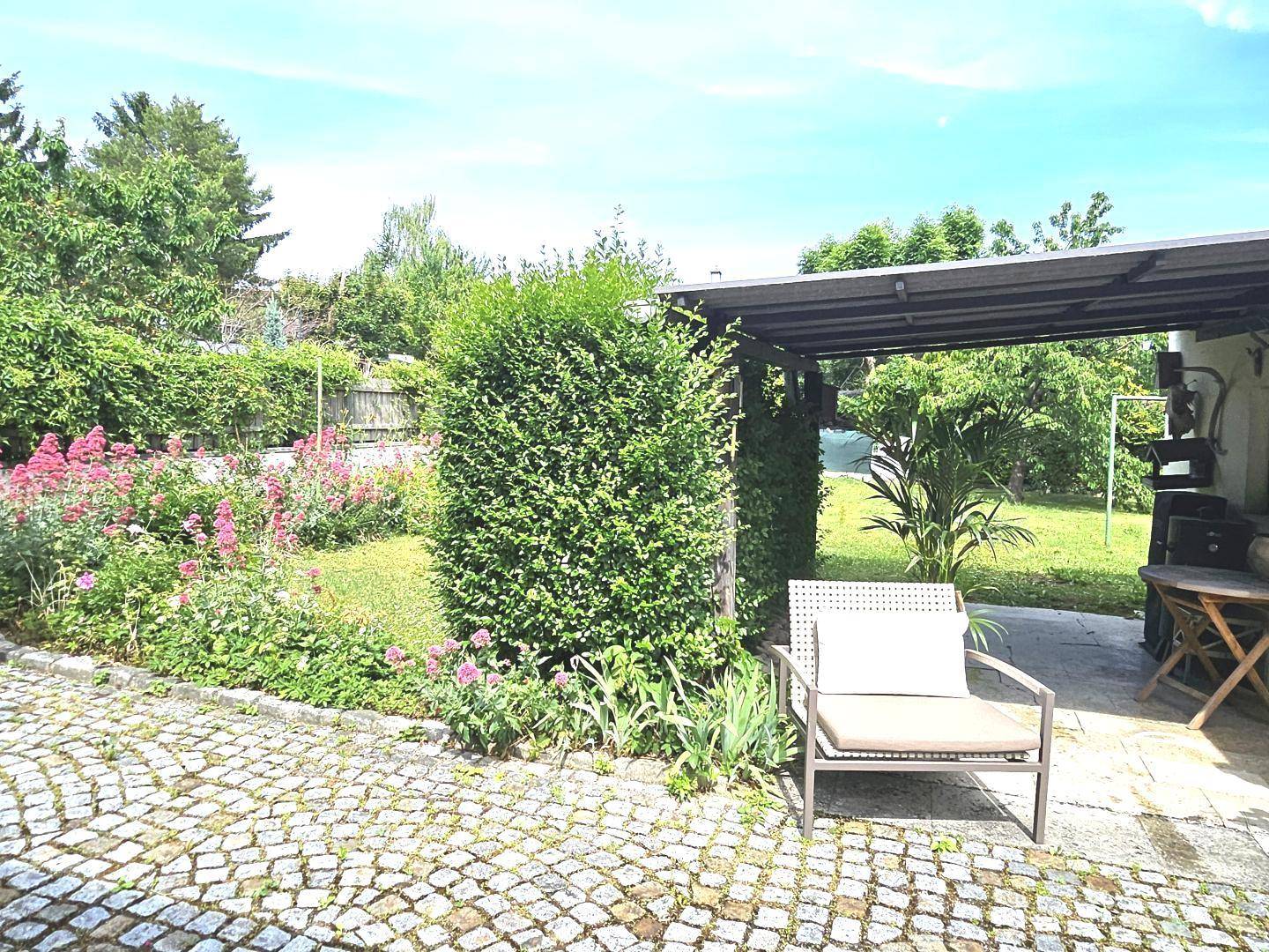 Gepflegter Gartenbereich mit Hecke, Blumenbeeten und einem überdachten Sitzplatz im Freien.