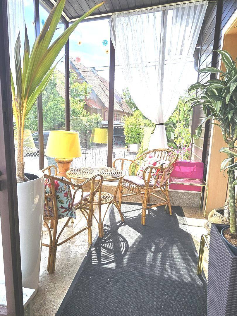 Heller Wintergarten mit Rattanmöbeln, vielen Pflanzen und großen Fenstern zum Garten.