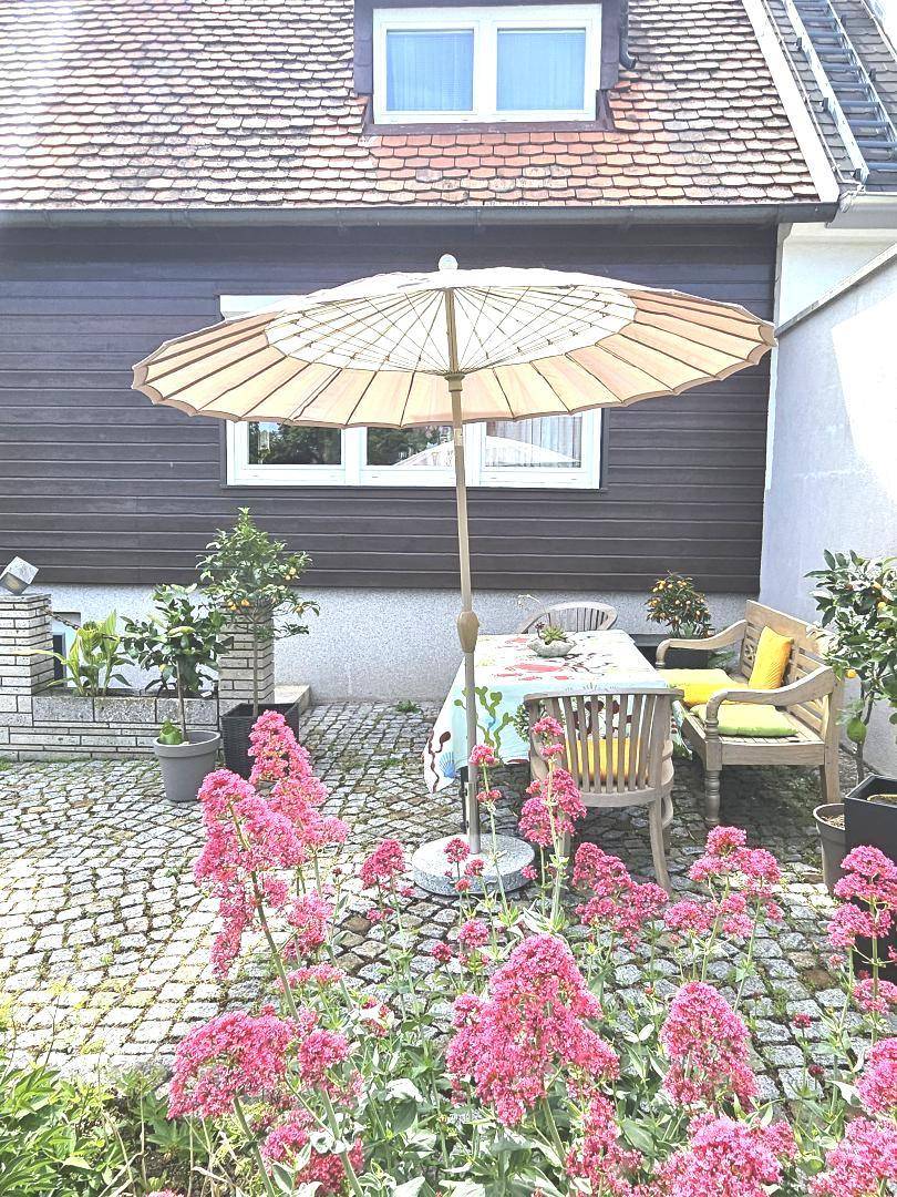 Gemütliche Terrasse mit Gartentisch, Sonnenschirm und blühenden Blumenbeeten vor der Hauswand.