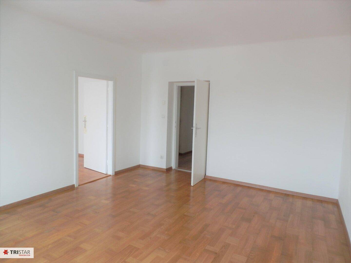 Geräumiges, leeres Zimmer mit hellem Parkettboden und zwei offenen Türen zu angrenzenden Räumen.