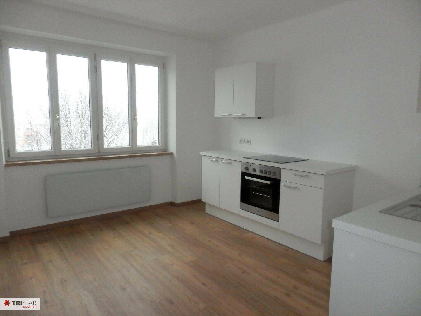 Einfache Küchenzeile mit Herd und Backofen in einem hellen Raum mit Parkettboden und großem Fenster.