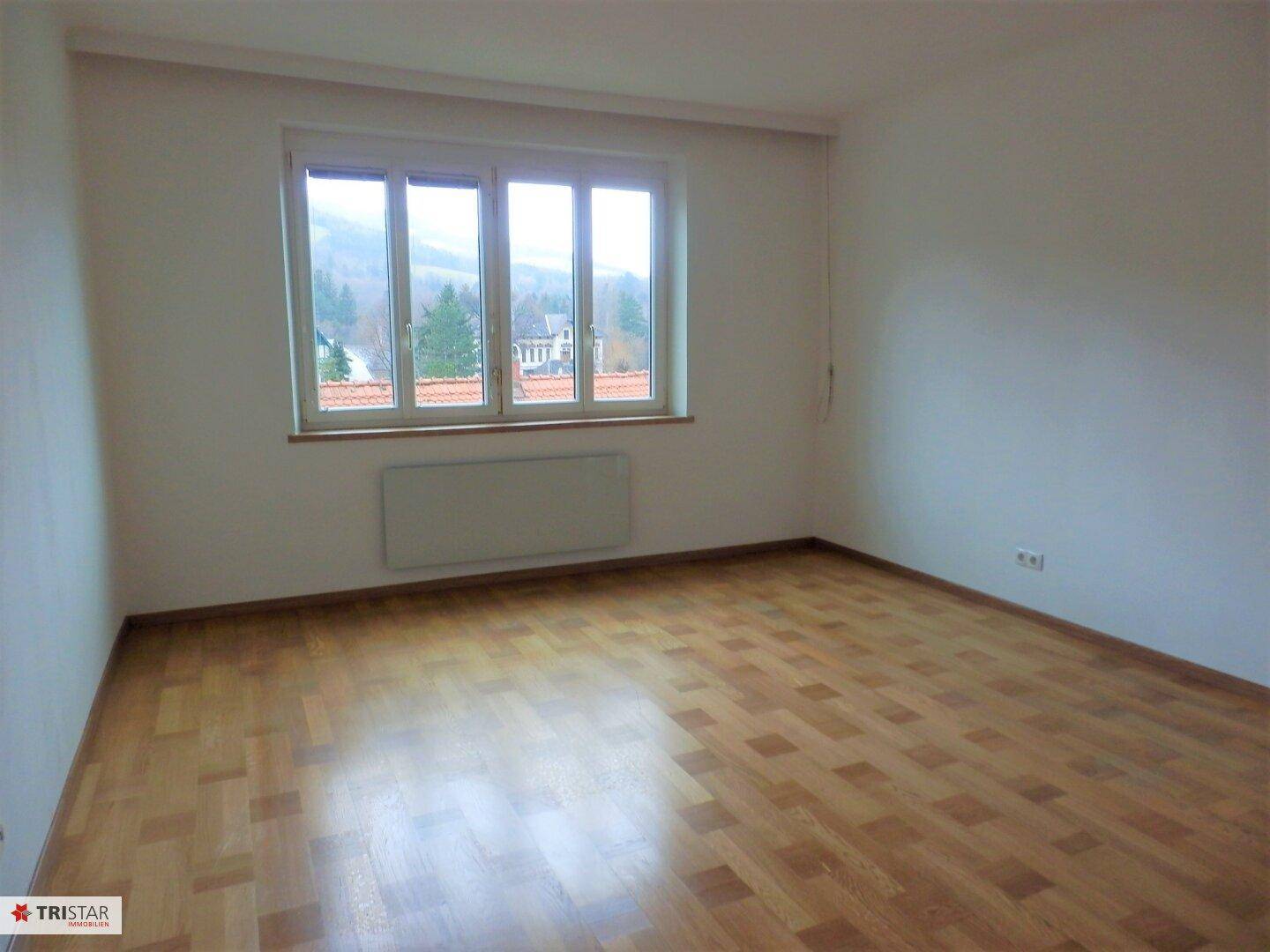 Helles, leeres Zimmer mit Parkettboden und großem Fenster mit Blick auf die grüne Umgebung.