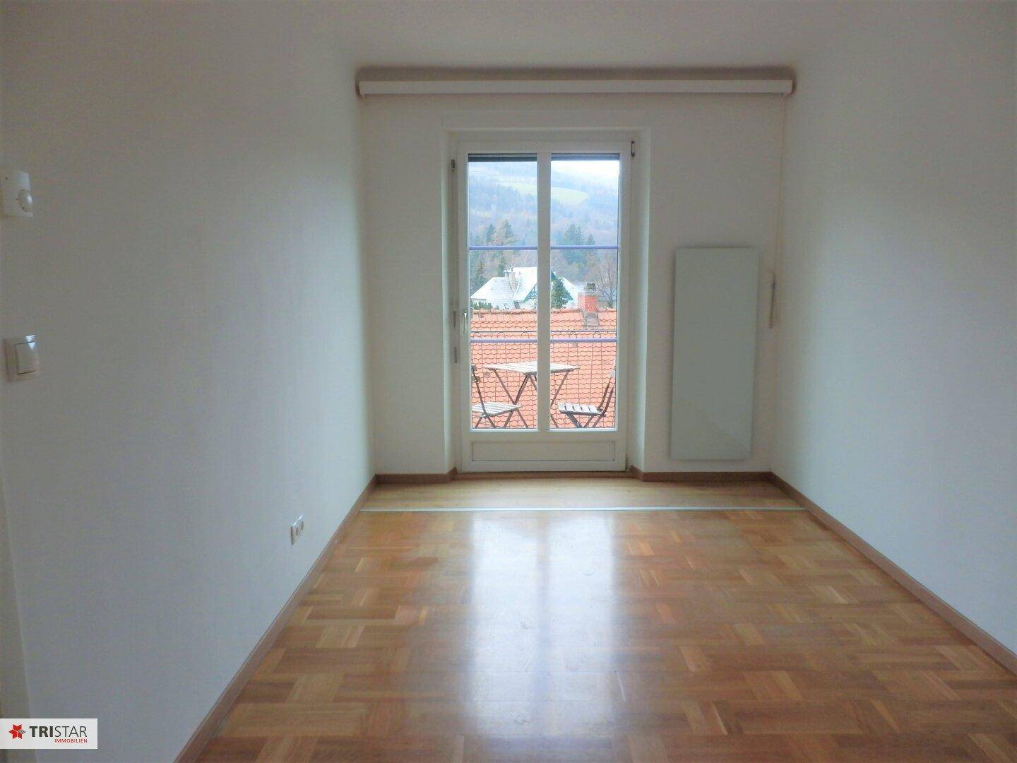 Helles Zimmer mit Parkettboden und Zugang zu einem kleinen Balkon mit Sitzgelegenheiten.