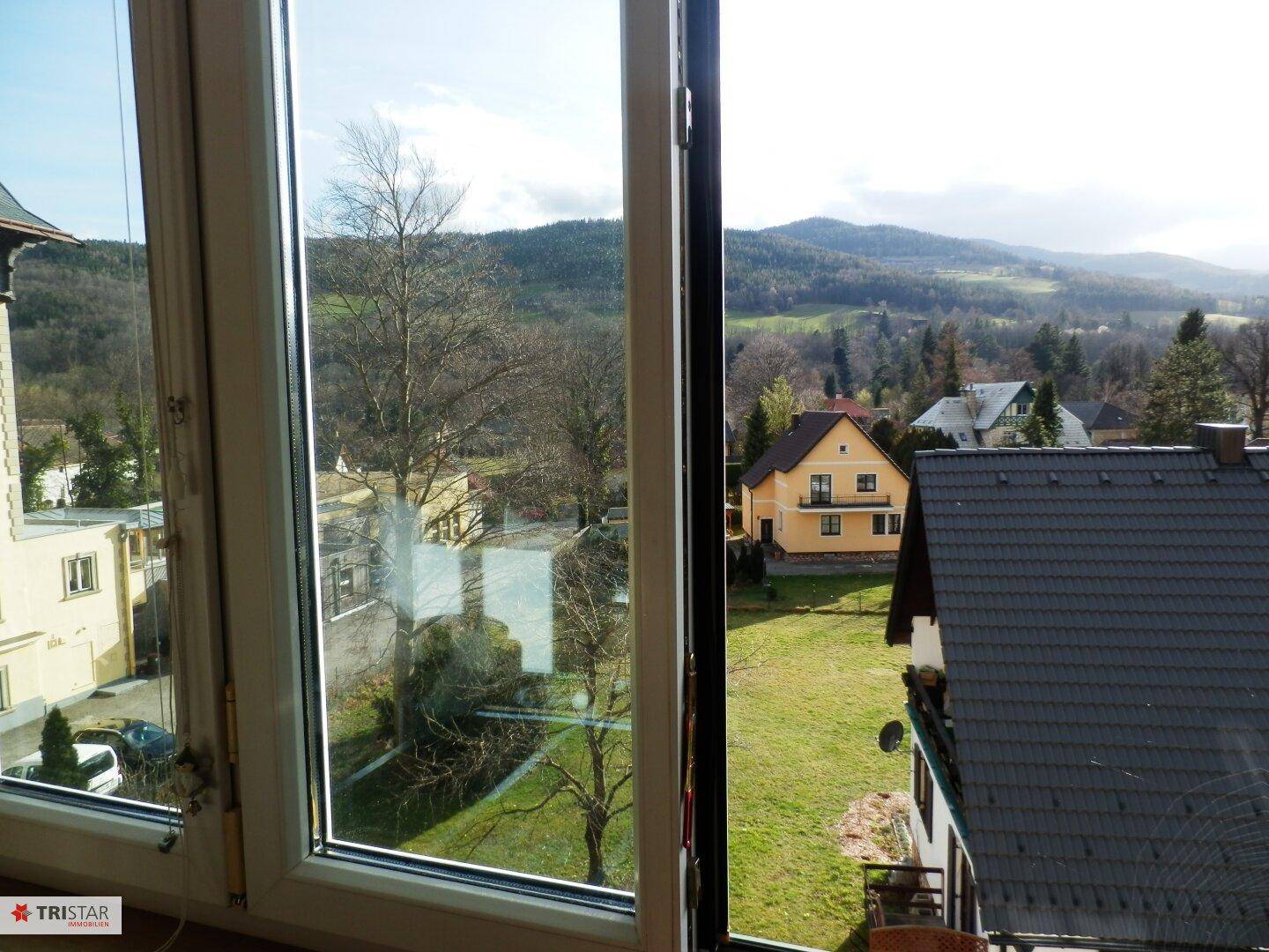 Ausblick aus einem geöffneten Fenster auf ein gelbes Haus, grüne Wiesen und bewaldete Hügel.