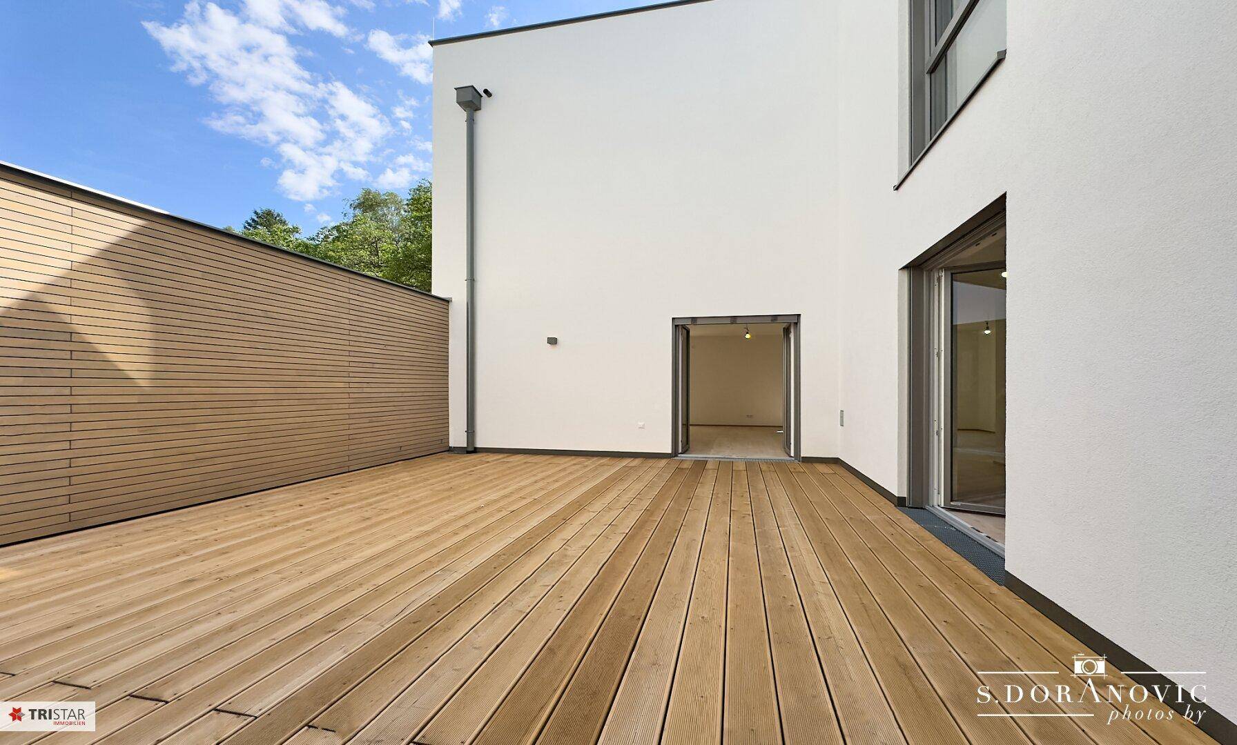 Sonniger Terrassenbereich mit Holzboden und direktem Zugang zu einem hellen Innenraum.