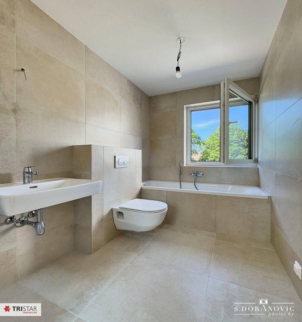 Modernes Badezimmer mit beigefarbenen Fliesen, Badewanne, WC und Fenster für Tageslicht.
