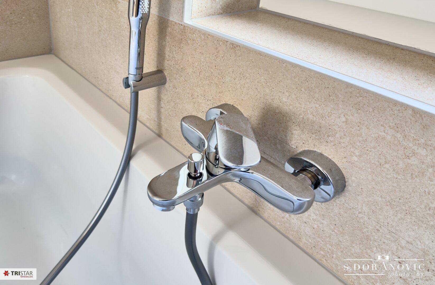 Detailaufnahme der Badewannenarmatur mit integrierter Handbrause und beigefarbenen Wandfliesen.