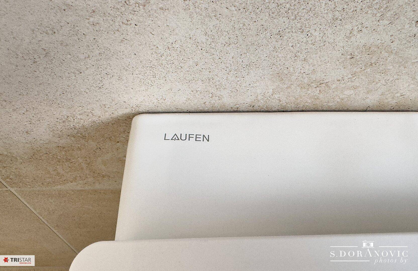 Nahaufnahme eines modernen Waschbeckens der Marke Laufen mit beigefarbenen Fliesen im Hintergrund.