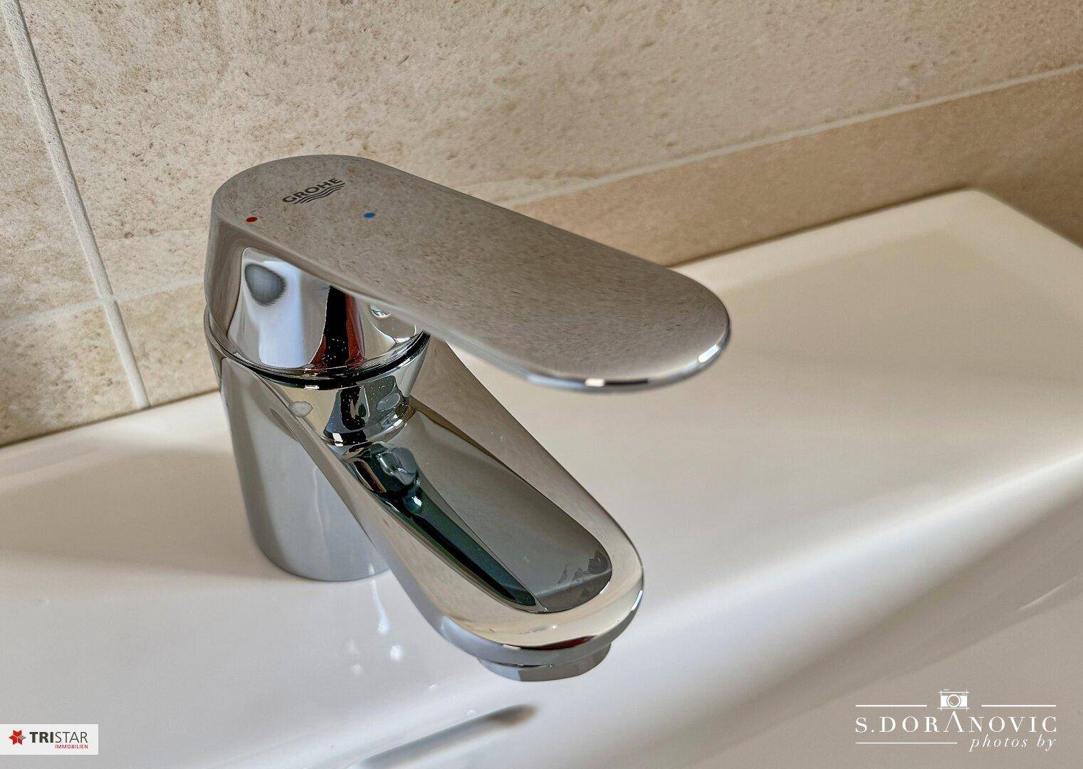 Detailansicht eines modernen Grohe-Wasserhahns auf einem weißen Waschbecken mit beigefarbenen Fliesen.