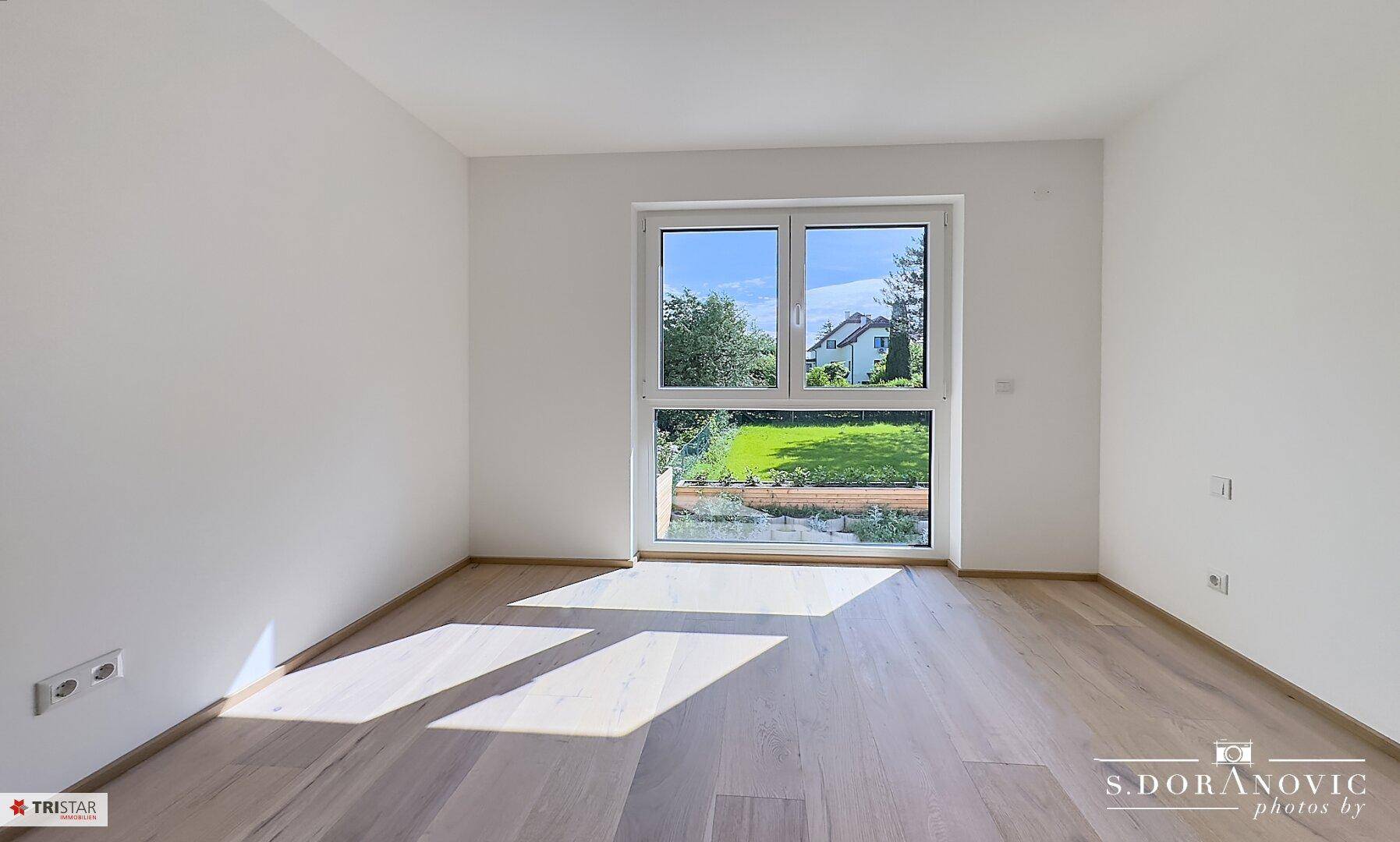 Heller, leerer Raum mit Holzboden und großem Fenster mit Blick ins Grüne.