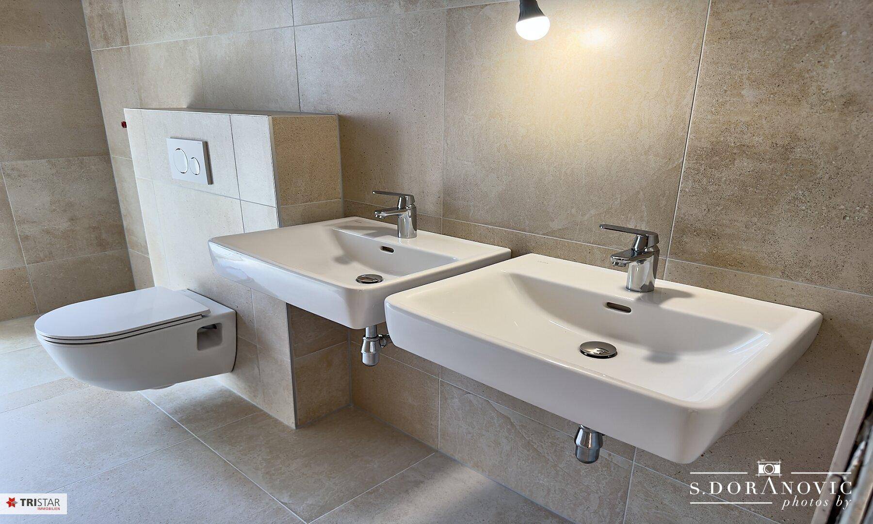 Helles Badezimmer mit zwei modernen Waschbecken, wandhängendem WC und beigefarbenen Fliesen.