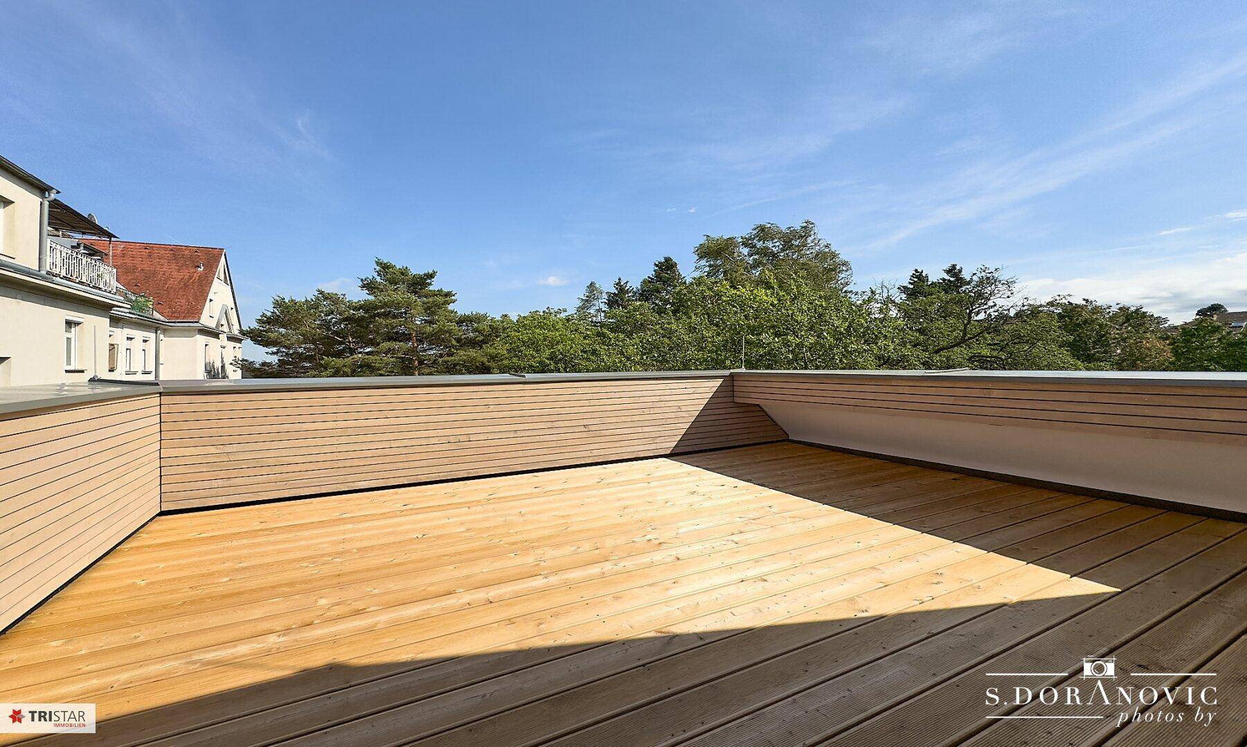 Weitläufige Dachterrasse mit hellem Holzdeck und freiem Blick auf den blauen Himmel und Bäume.