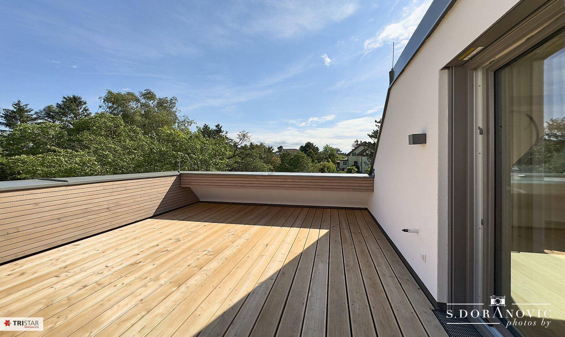 Sonnige Dachterrasse mit Holzbelag und Zugang zum Innenbereich, umgeben von grünen Bäumen.
