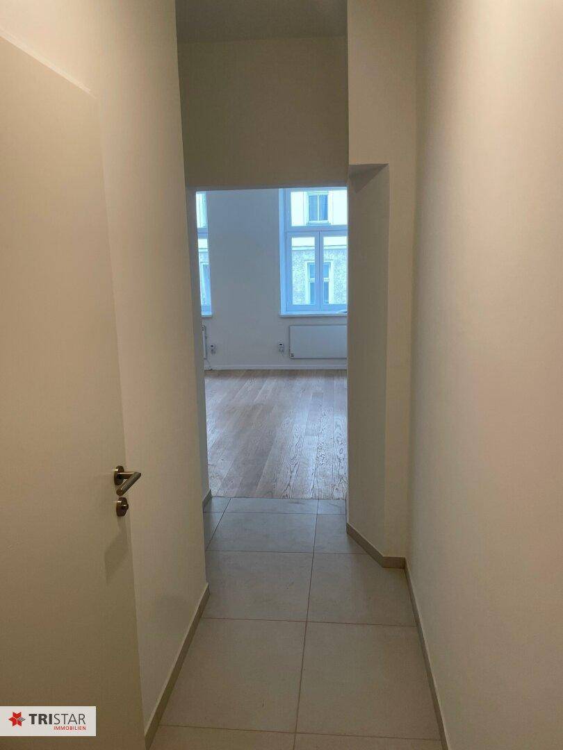 Heller Flur mit Fliesenboden, der zu einem lichtdurchfluteten Raum mit Parkett und Fenstern führt.