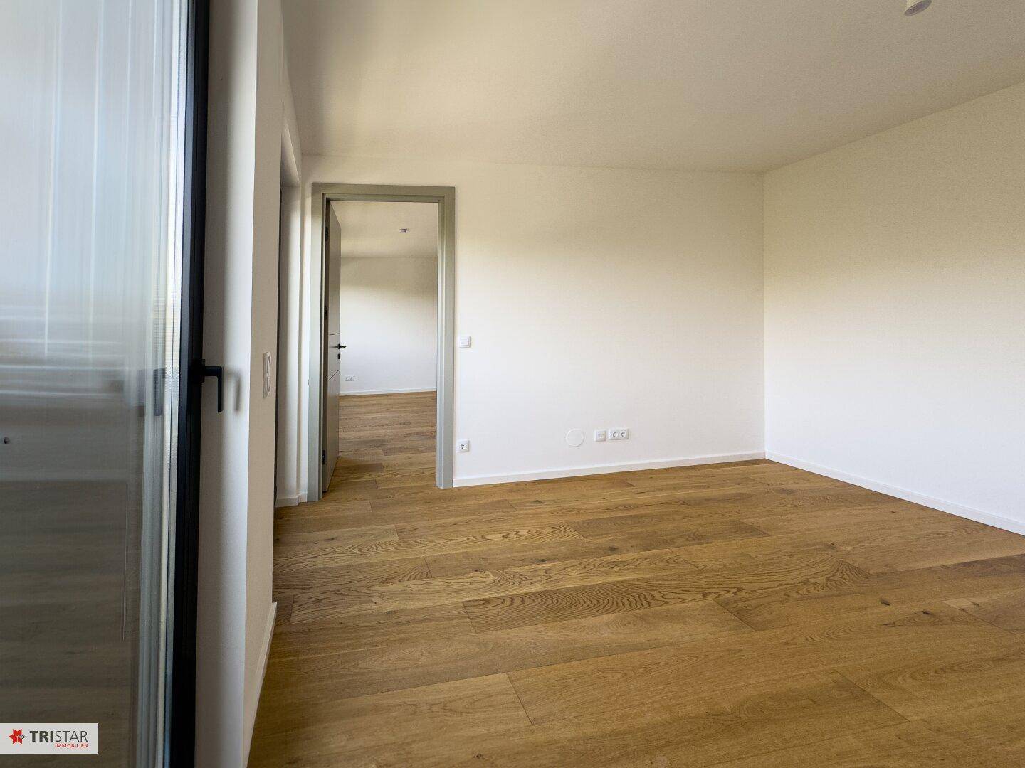 Heller Raum mit Holzboden und Blick in den Flur, der weitere Zimmer verbindet.