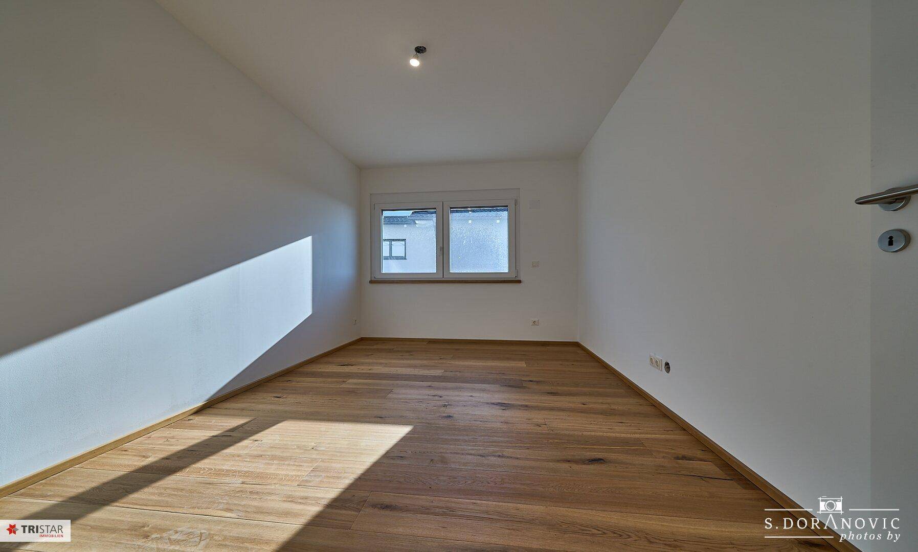 Leerer, heller Raum mit Holzboden und einem Fenster, das für eine angenehme Belichtung sorgt.