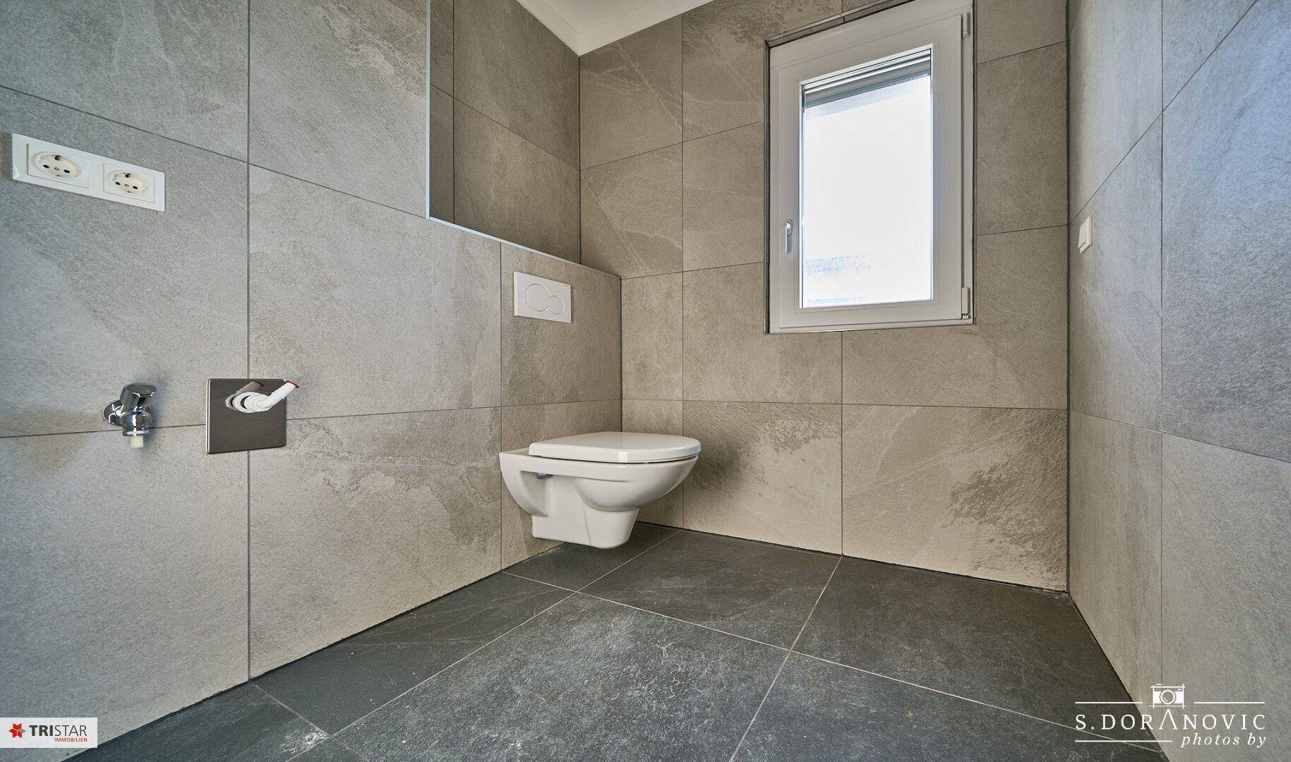 Modernes WC mit wandhängender Toilette und Fenster, komplett in Grautönen gefliest.