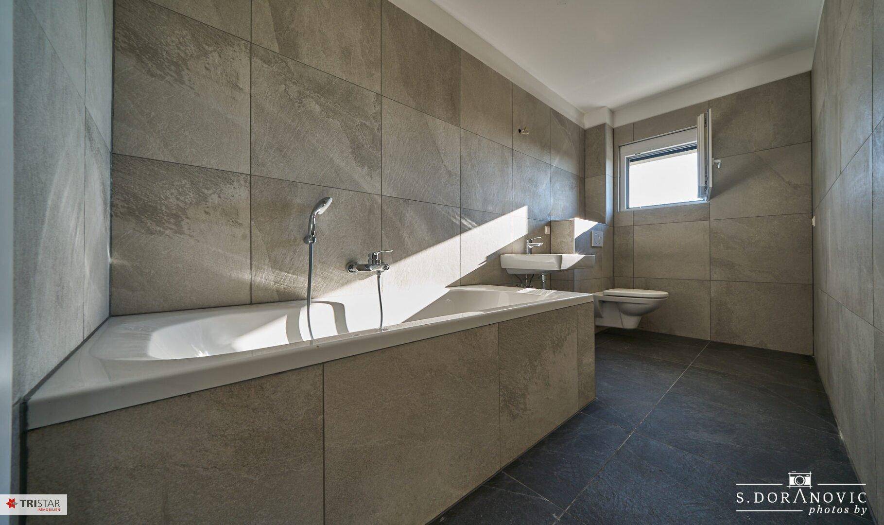 Geräumiges Badezimmer mit Badewanne, Toilette und Fenster, komplett gefliest.