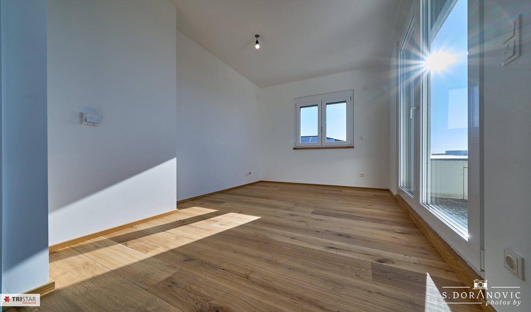 Geräumiger, heller Raum mit Holzboden, Fenster und großer Glastür, die nach draußen führt.