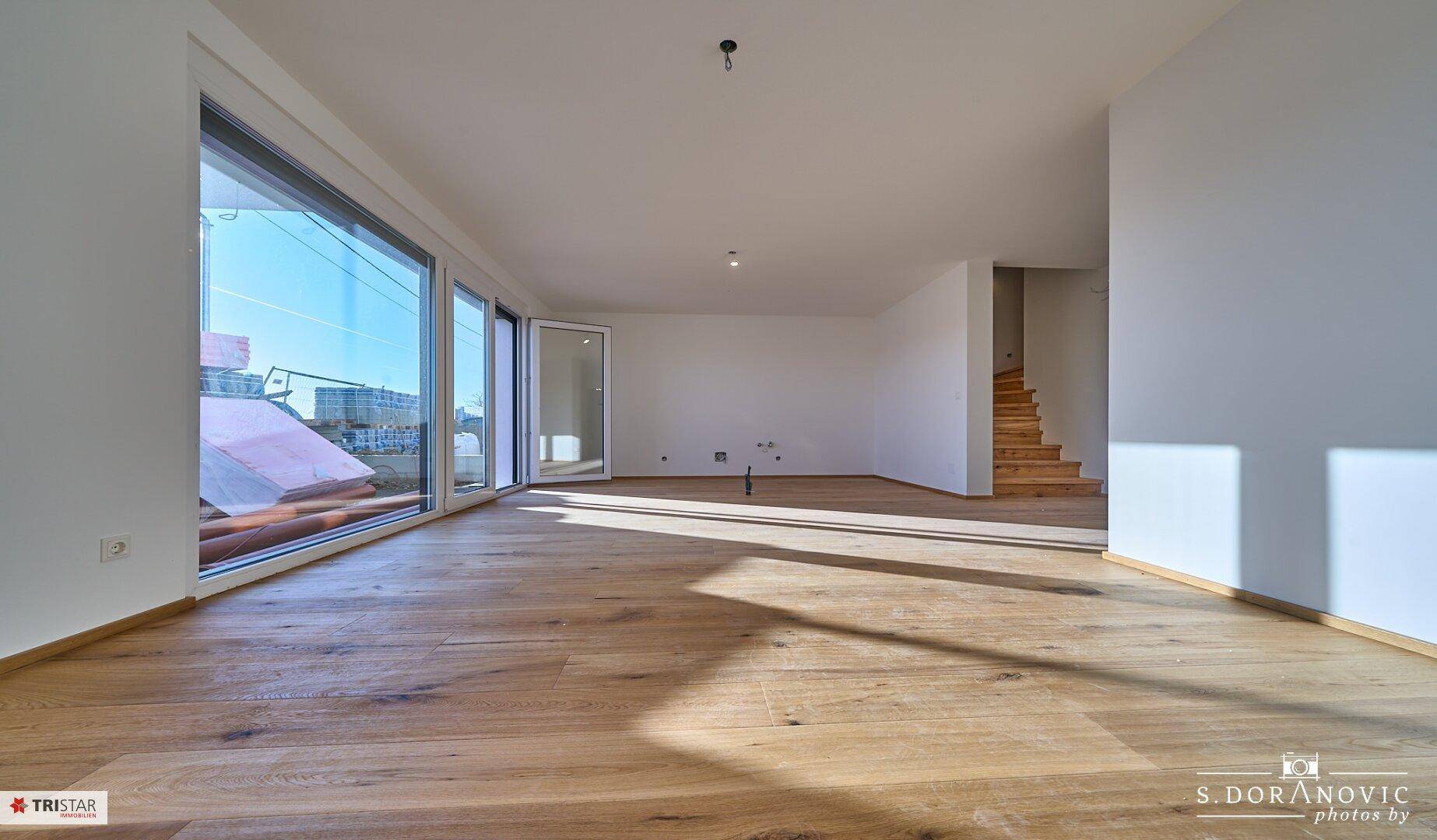 Geräumiger Wohnbereich mit Holzboden, großen Fenstern und Blick auf eine Treppe.