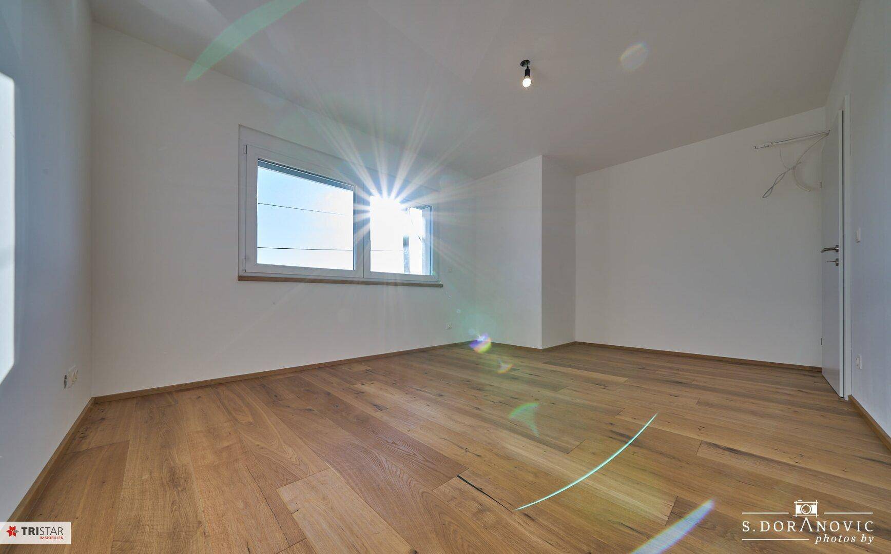 Heller, leerer Raum mit Holzboden und einem Fenster, das viel Sonnenlicht hereinlässt.