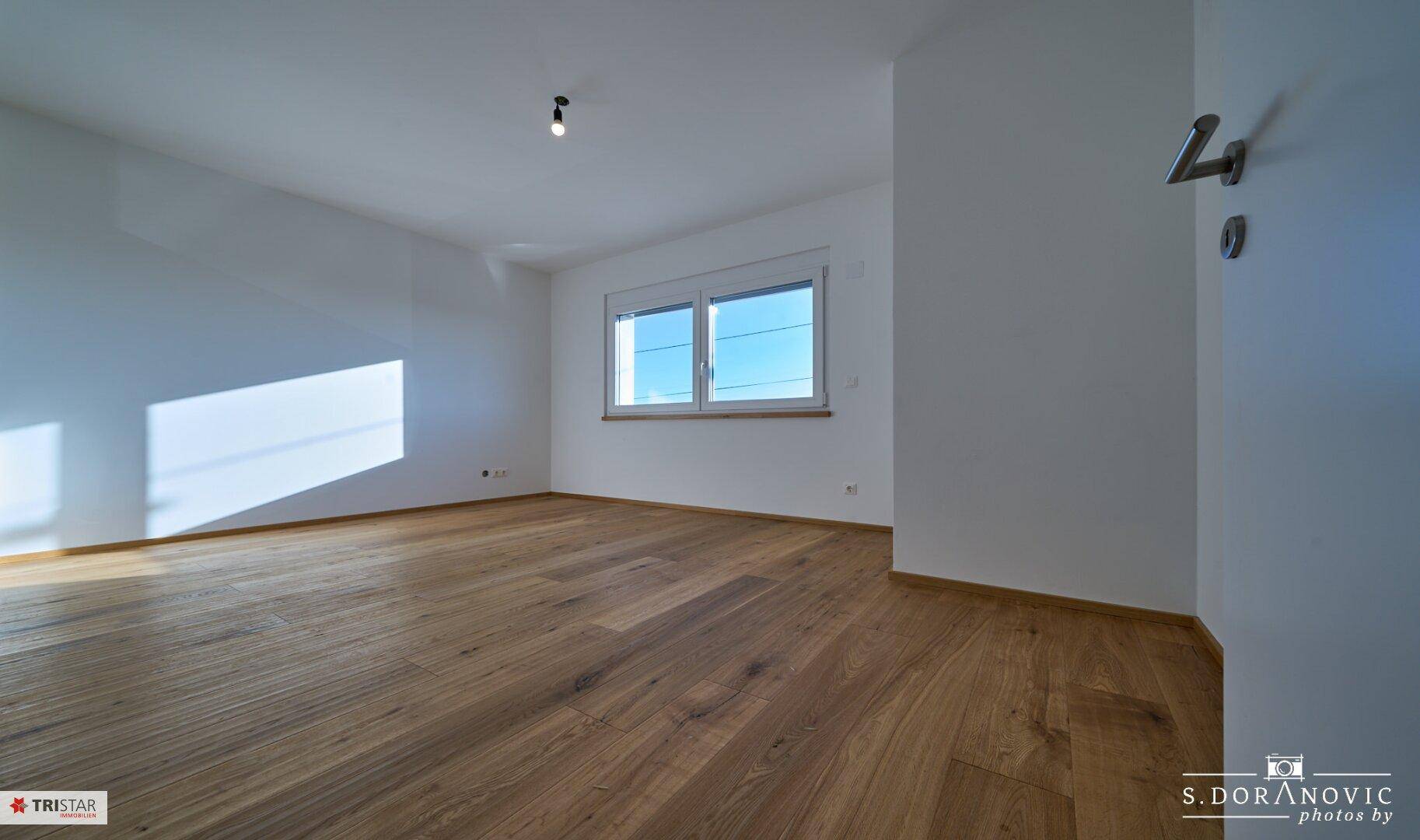 Helles Zimmer mit Holzboden und großen Fenstern, das viel Tageslicht hereinlässt.
