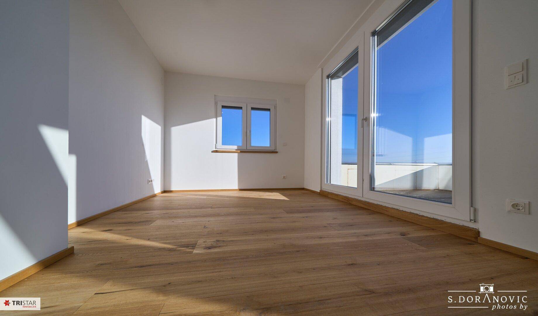Geräumiges Zimmer mit Holzboden und großen Fenstern, die auf eine Terrasse führen.