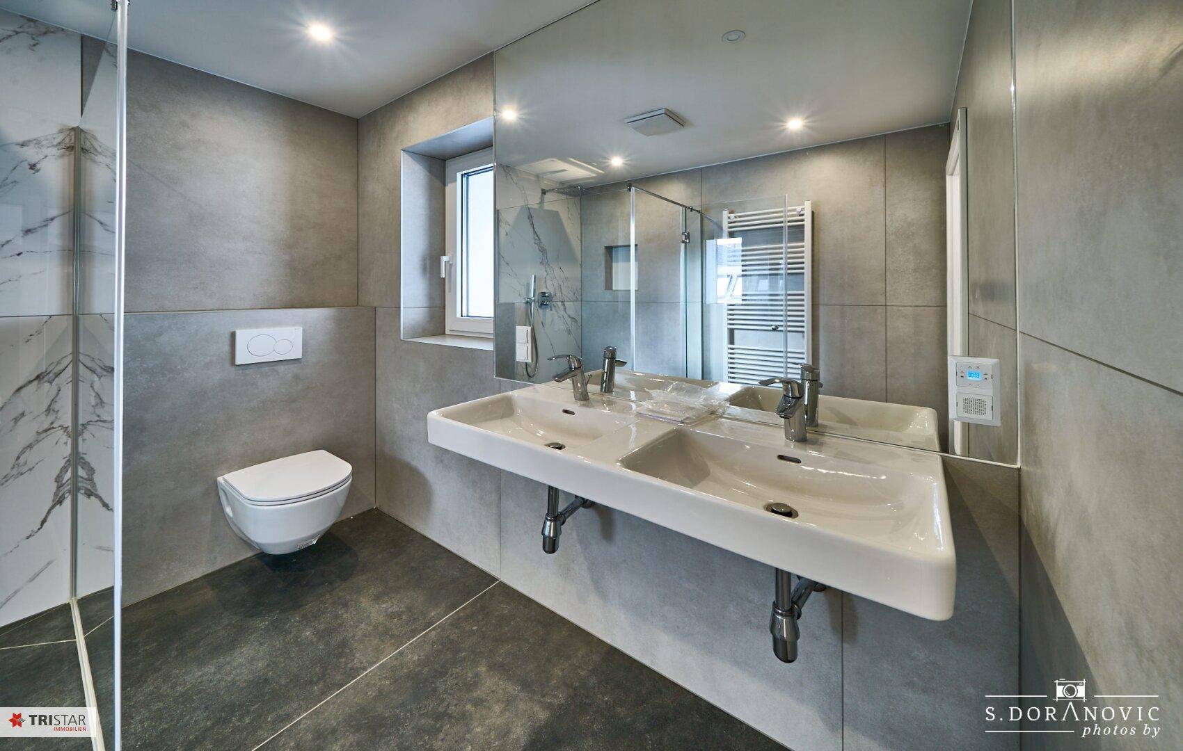 Modernes Badezimmer mit Doppelwaschbecken, begehbarer Dusche und Fenster, in Grautönen gehalten.