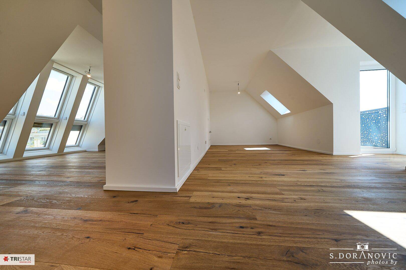 Großzügiger Dachgeschossraum mit Holzboden, schrägen Wänden und verschiedenen Fenstern für viel Licht.