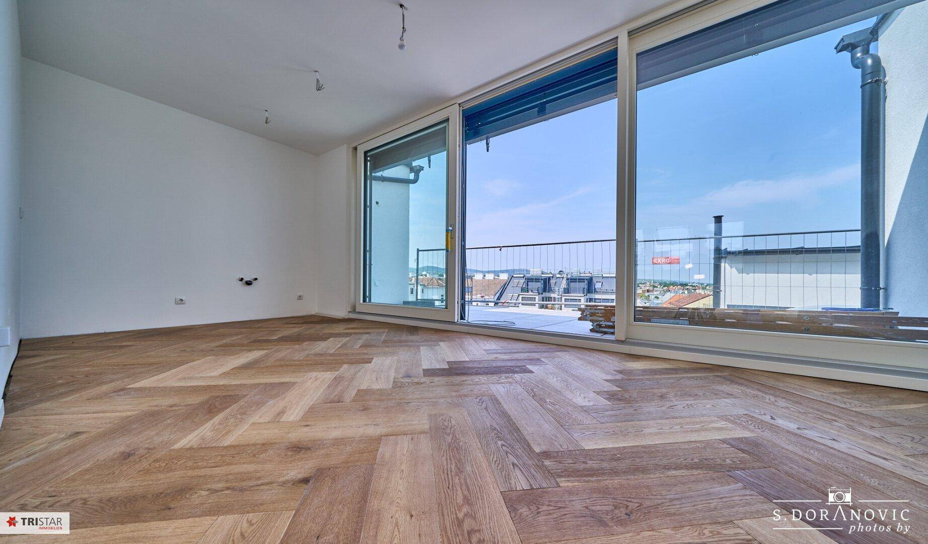 Lichtdurchfluteter Raum mit Fischgrätparkett und breiter Schiebetür zur Dachterrasse mit Stadtblick.