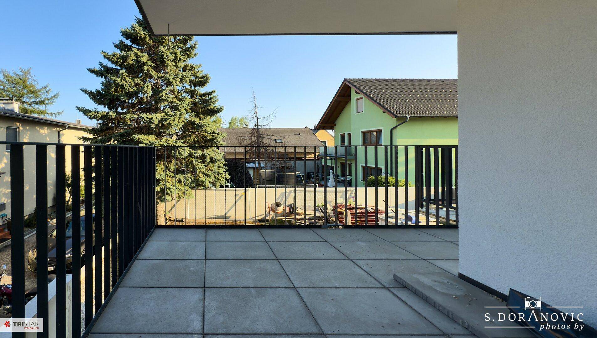 Heller Balkon mit modernem Fliesenboden und schwarzem Geländer, bietet einen Blick auf die umliegenden Häuser und Bäume.