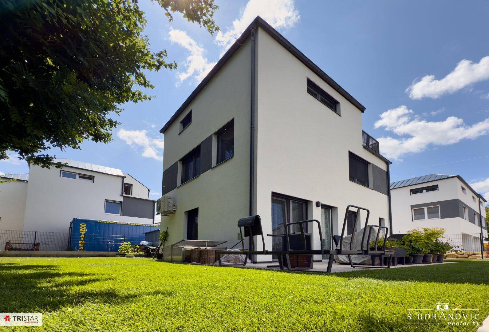 Modernes Haus mit weißer Fassade, grünem Rasen und Außensitzbereich unter blauem Himmel.