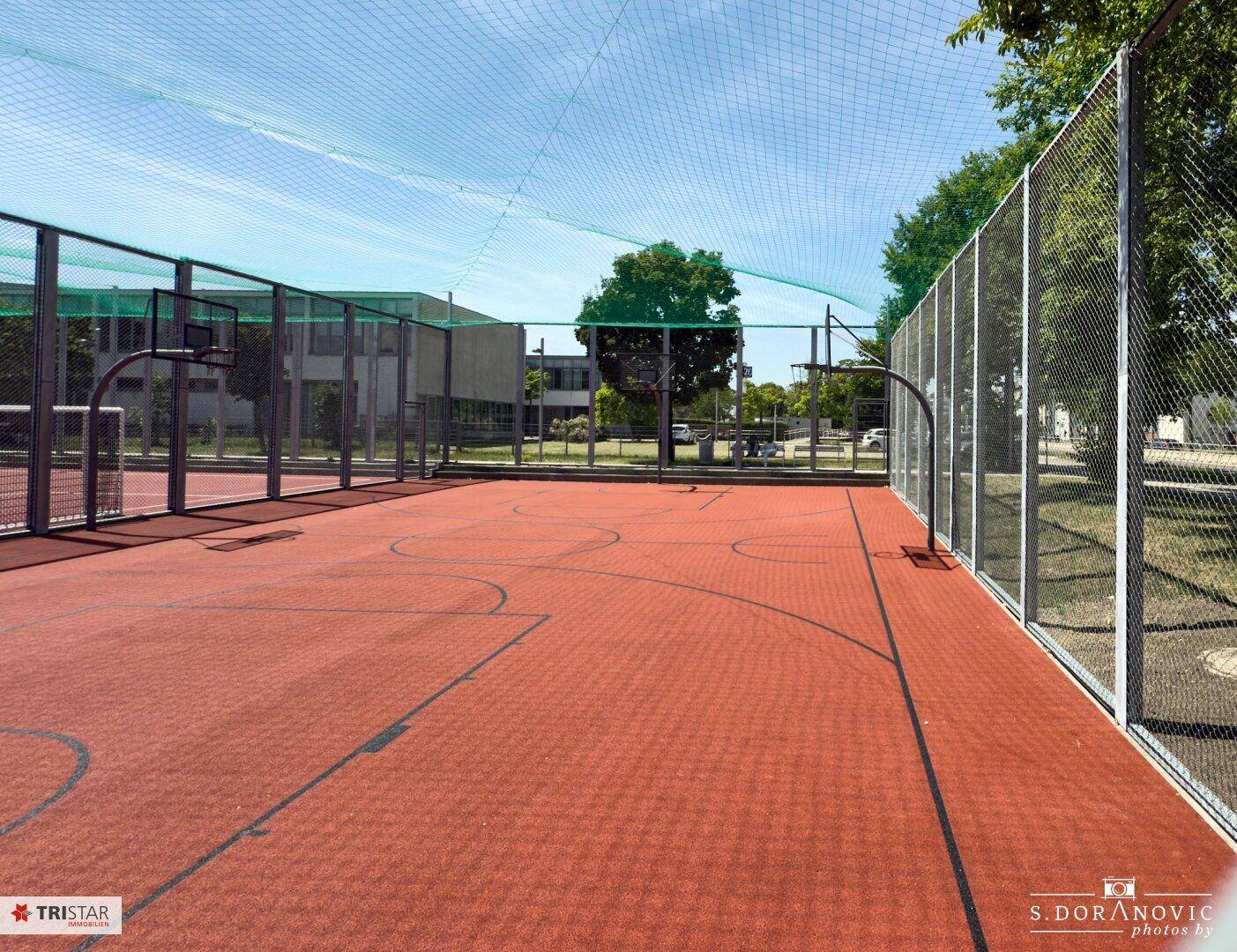 Ein roter Multisportplatz mit Basketballkorb und hohen Zäunen unter blauem Himmel.