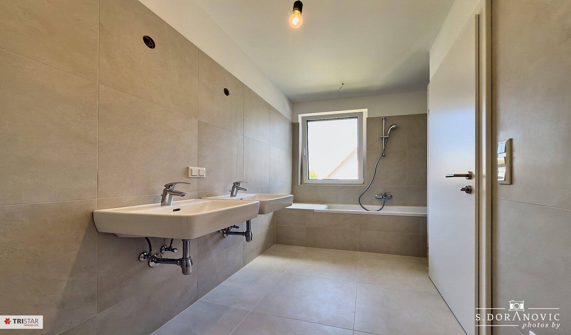 Modernes Badezimmer mit hellgrauen Fliesen, Doppelwaschbecken und einer Badewanne mit Duschfunktion.