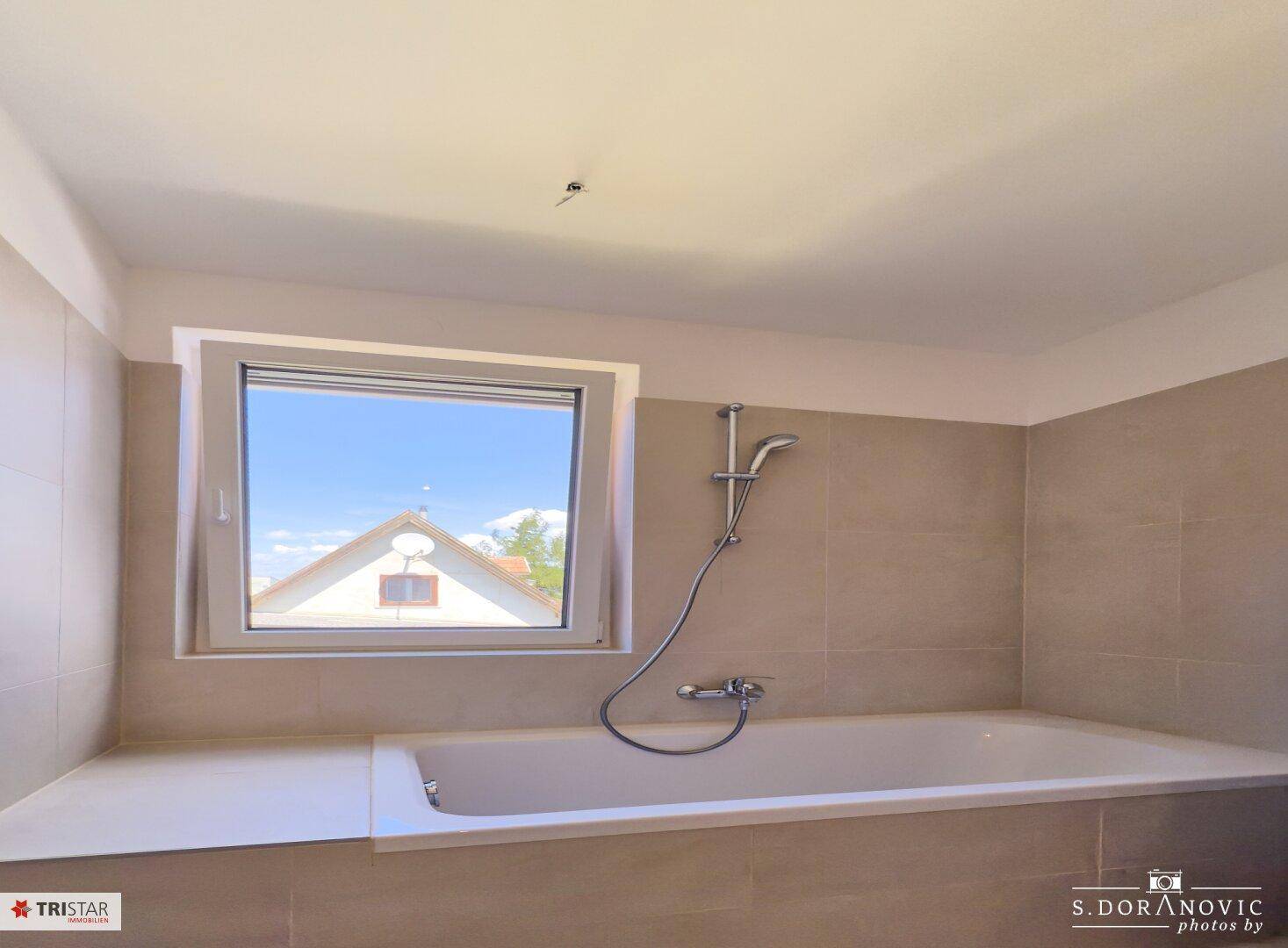 Badezimmer mit Badewanne und Fenster, ausgestattet mit hellgrauen Wand- und Bodenfliesen.