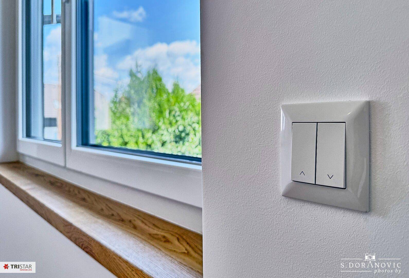 Nahaufnahme eines Lichtschalters an einer weißen Wand mit Blick ins Grüne durch das Fenster.