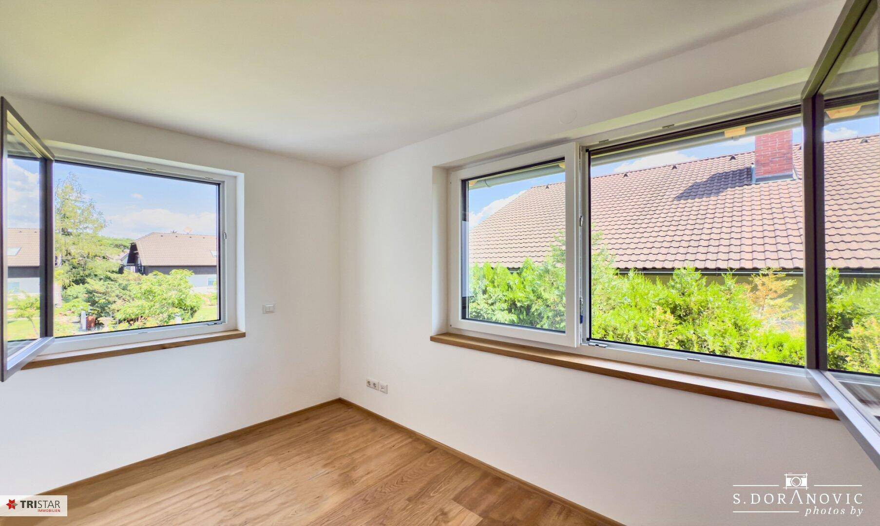 Helles, leeres Zimmer mit Holzboden und großen Fenstern mit Blick ins Grüne.
