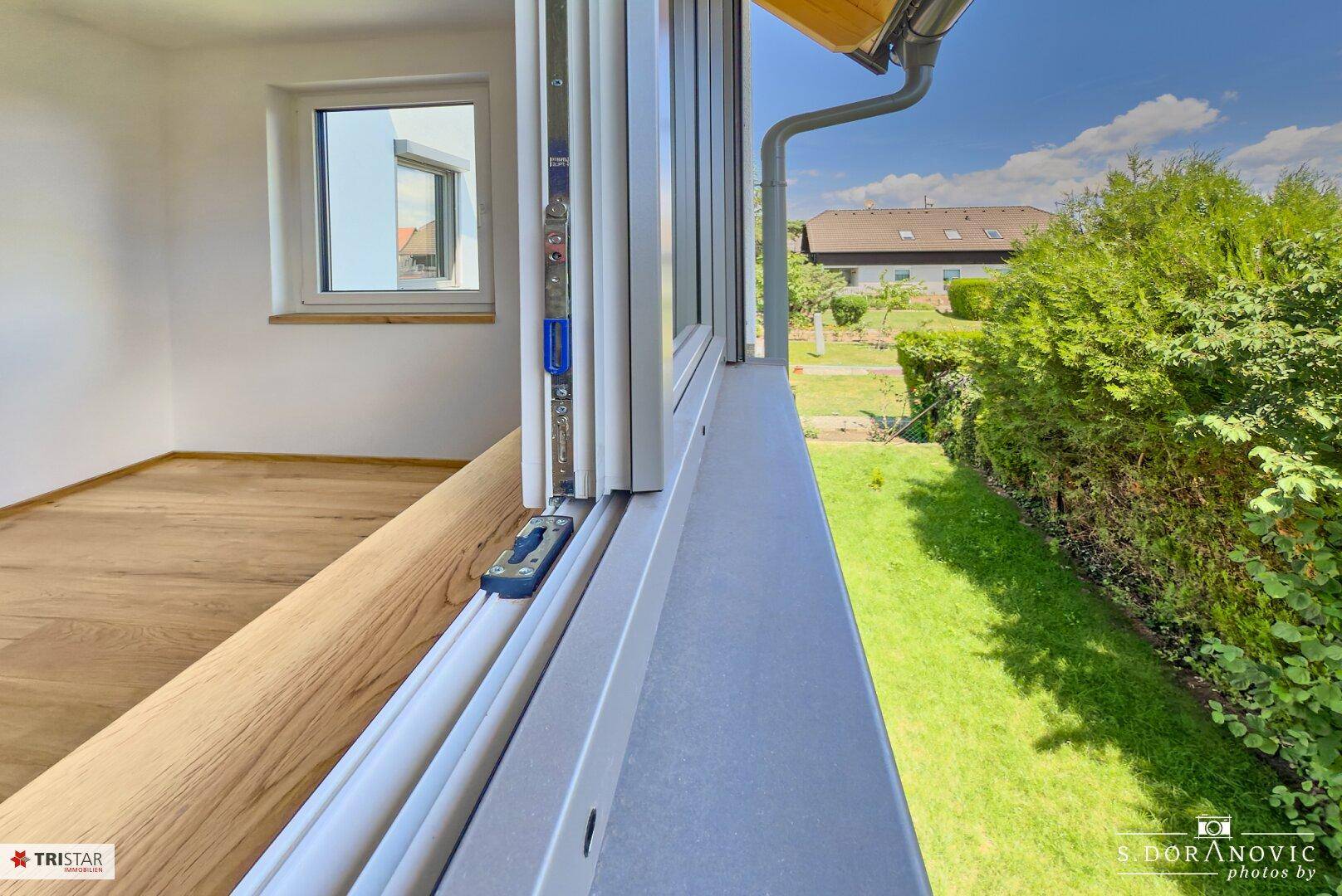 Fensterblick auf den Garten mit Rasenfläche und Hecken unter blauem Himmel.