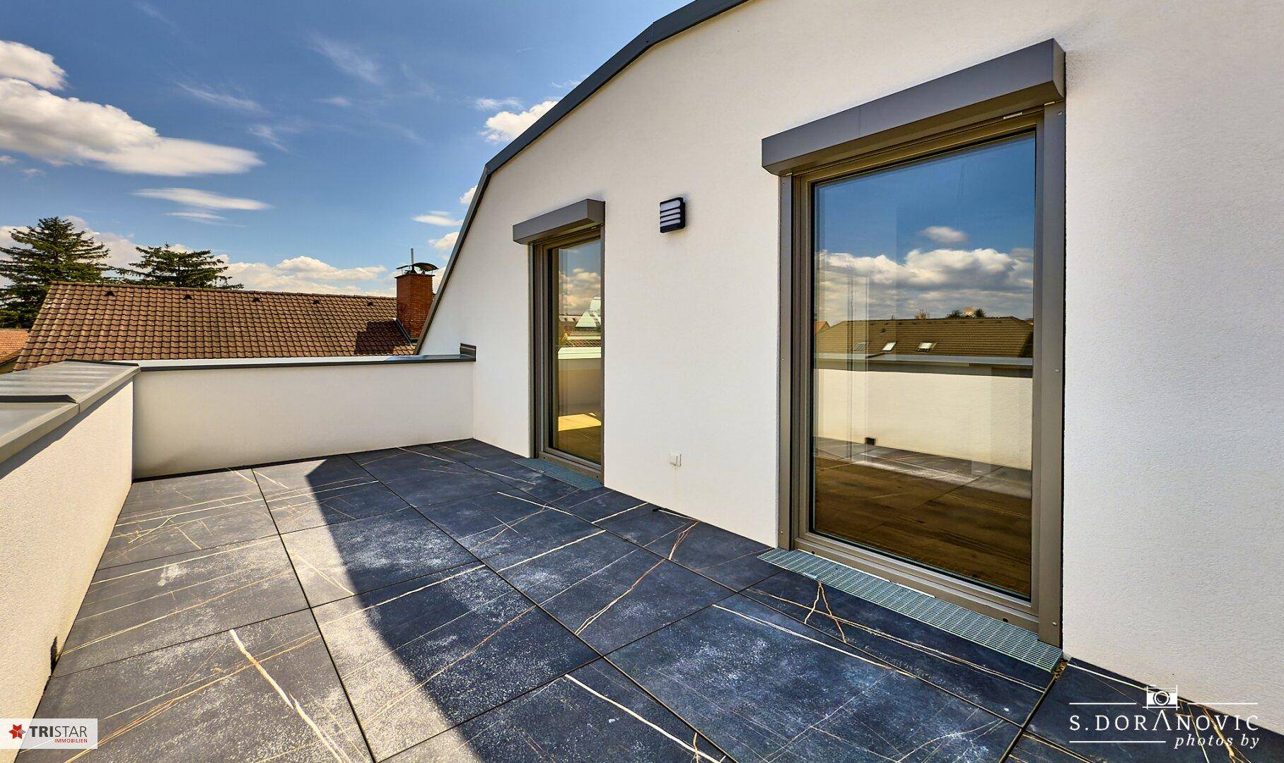 Geräumige Dachterrasse mit modernen Fliesen und großen Fenstertüren unter blauem Himmel.
